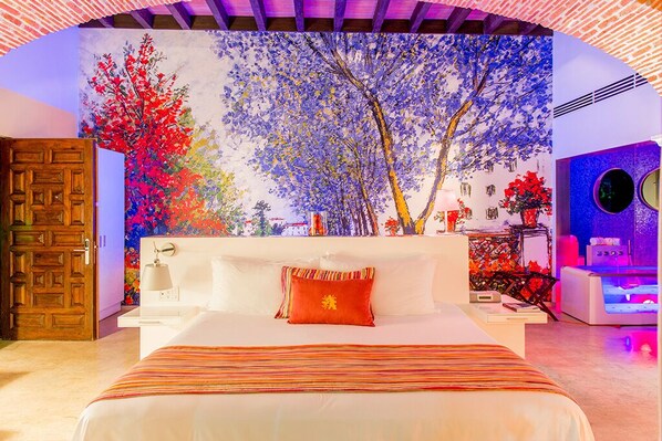 A hotel room with a large mural of trees, a bed with a striped bedspread, a wooden door, and a small table with a lamp.