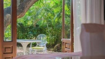 Quarto com uma janela ampla que dá para um jardim exuberante. Há uma porta de madeira, uma mesa branca e uma cadeira de plástico transparente.