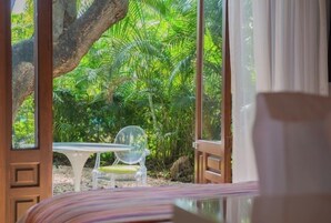 Habitación con un ventanal grande que da a un jardín frondoso. Se ve una puerta de madera, una mesa blanca y una silla de plástico transparente.