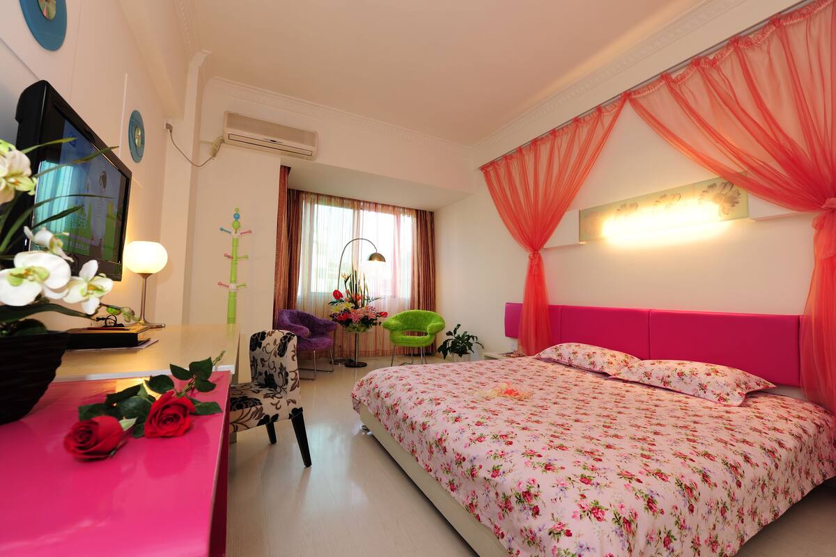 A bedroom with a floral bedspread, a pink headboard, a desk with a lamp, and a chair.