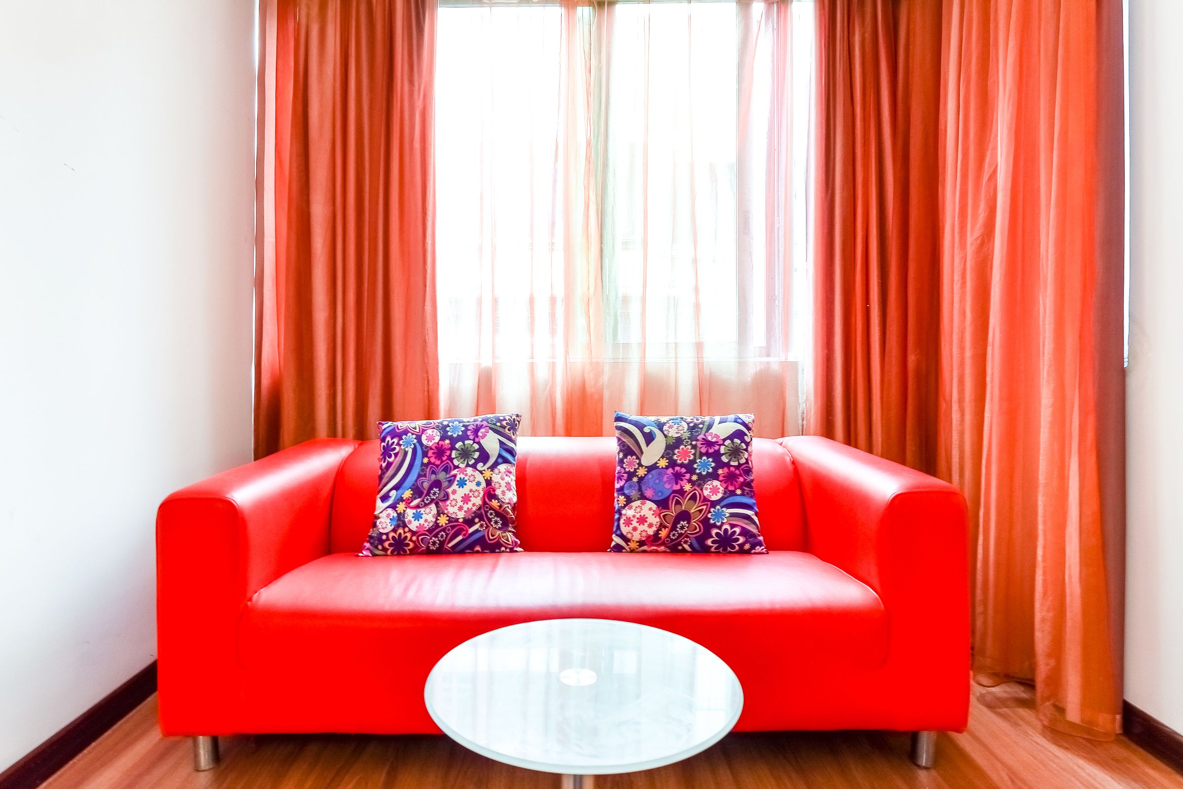 A red sofa with floral cushions, a glass coffee table, and orange curtains.