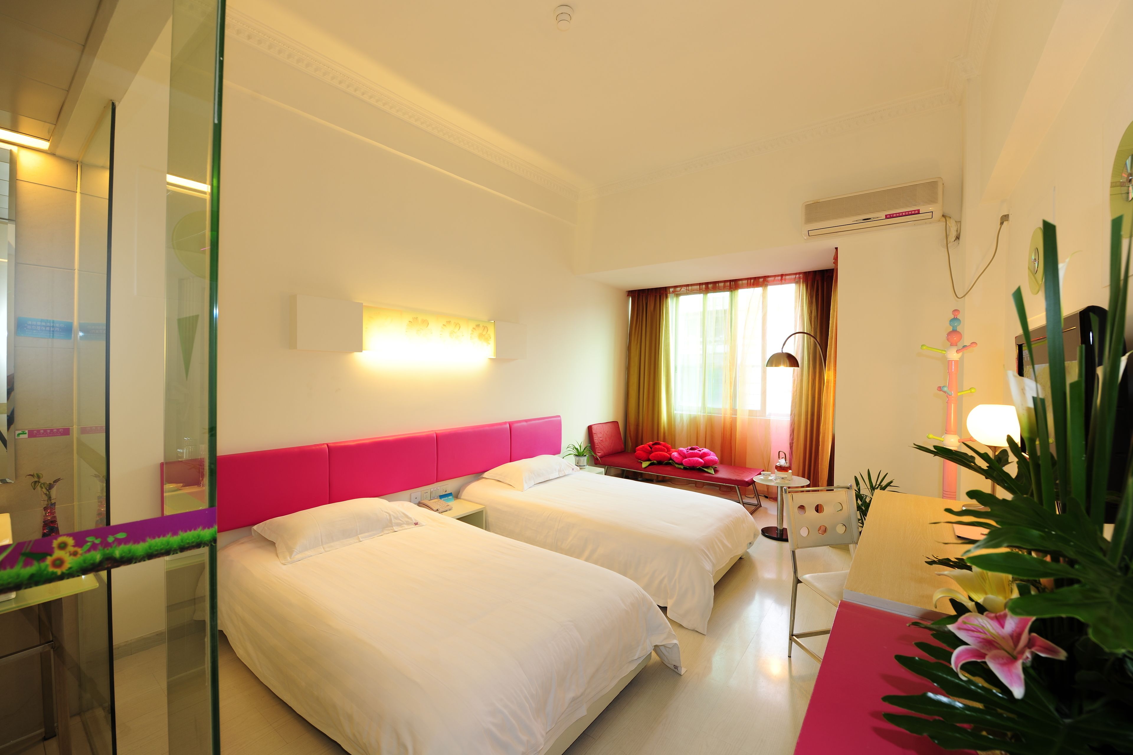 A hotel room with two beds, a glass partition, a red headboard, a desk with a chair, a window with curtains, and a potted plant.