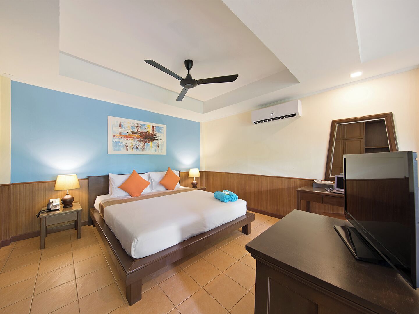 Quarto de hotel com cama, mesas de cabeceira, escrivaninha, ventoinha no teto e um ar condicionado, além de um quadro na parede.