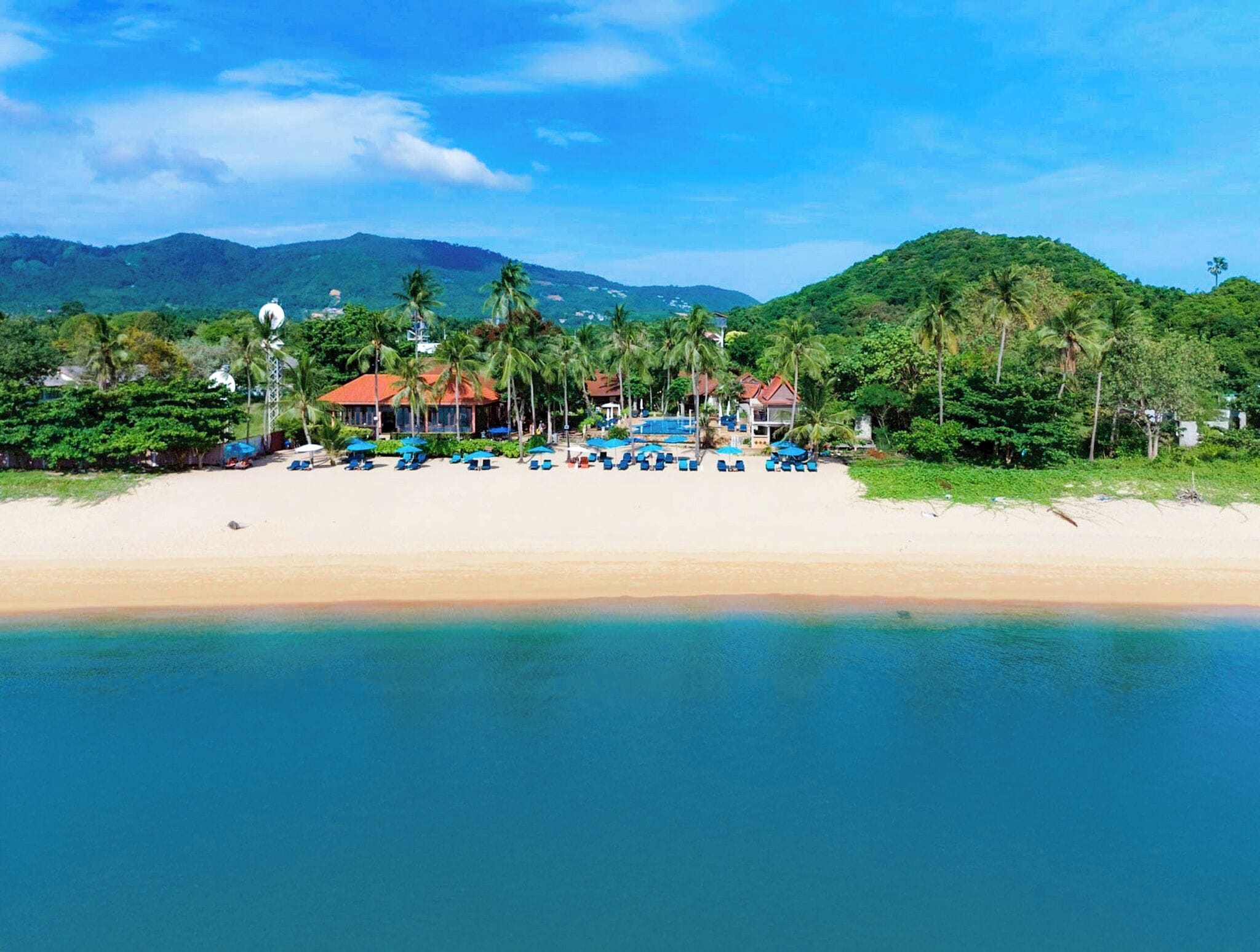Ein Strandresort mit mehreren bungalowartigen Gebäuden, einem Sandstrand und Palmen.