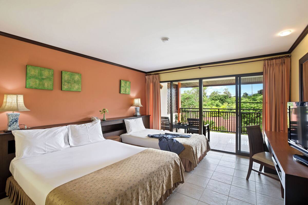 A hotel room with two beds, a TV, a desk, and a balcony with a view of greenery.