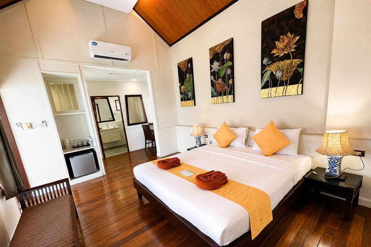 A hotel room with a wooden floor, a bed with white bedding and orange pillows, a nightstand with a lamp, and artwork on the wall.