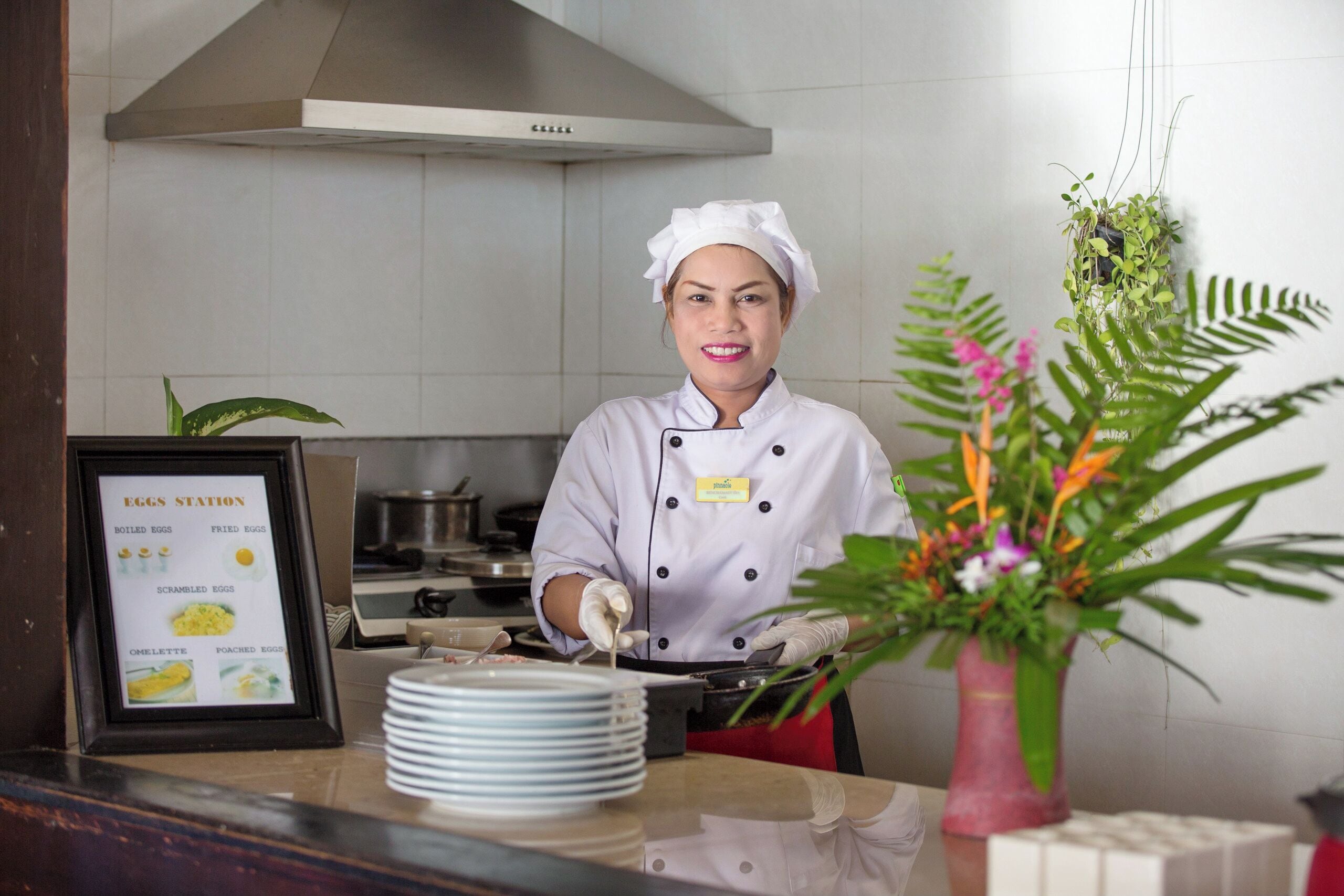 Un chef con uniforme blanco y una tarjeta de identificación, preparando comida en una barra con un panel de menú que dice 'EGGS STATION' y enumera varios platos de huevo.