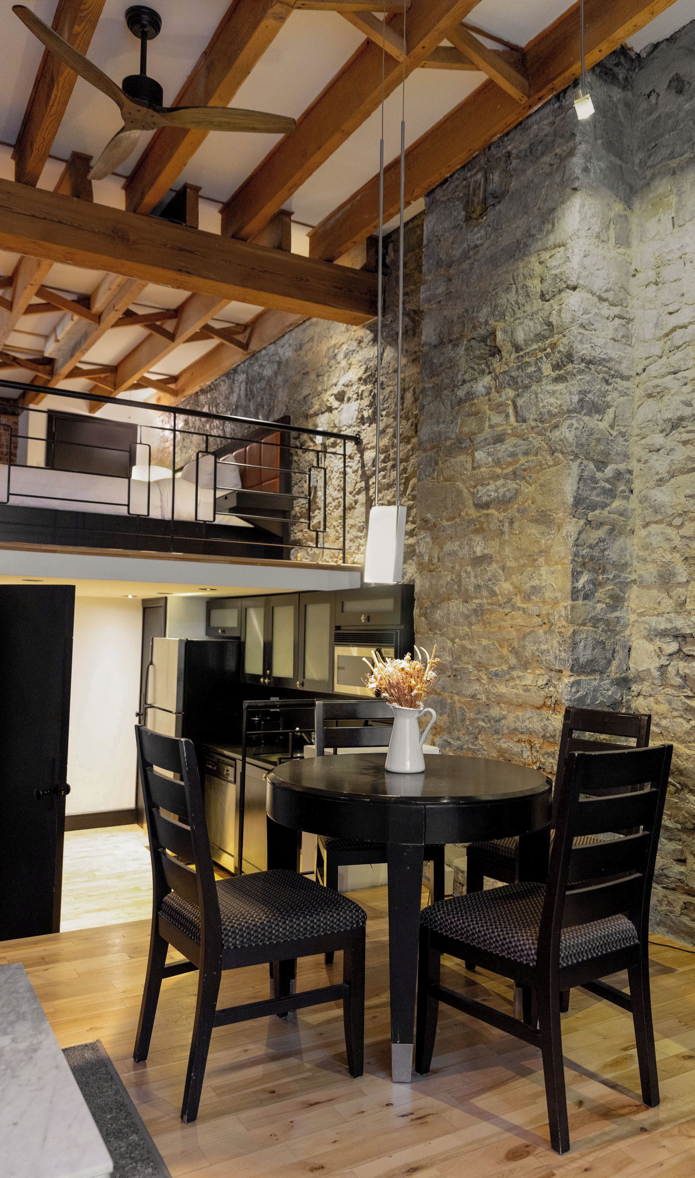 A dining area with a round table and four chairs, a kitchen in the background, and a stone wall.