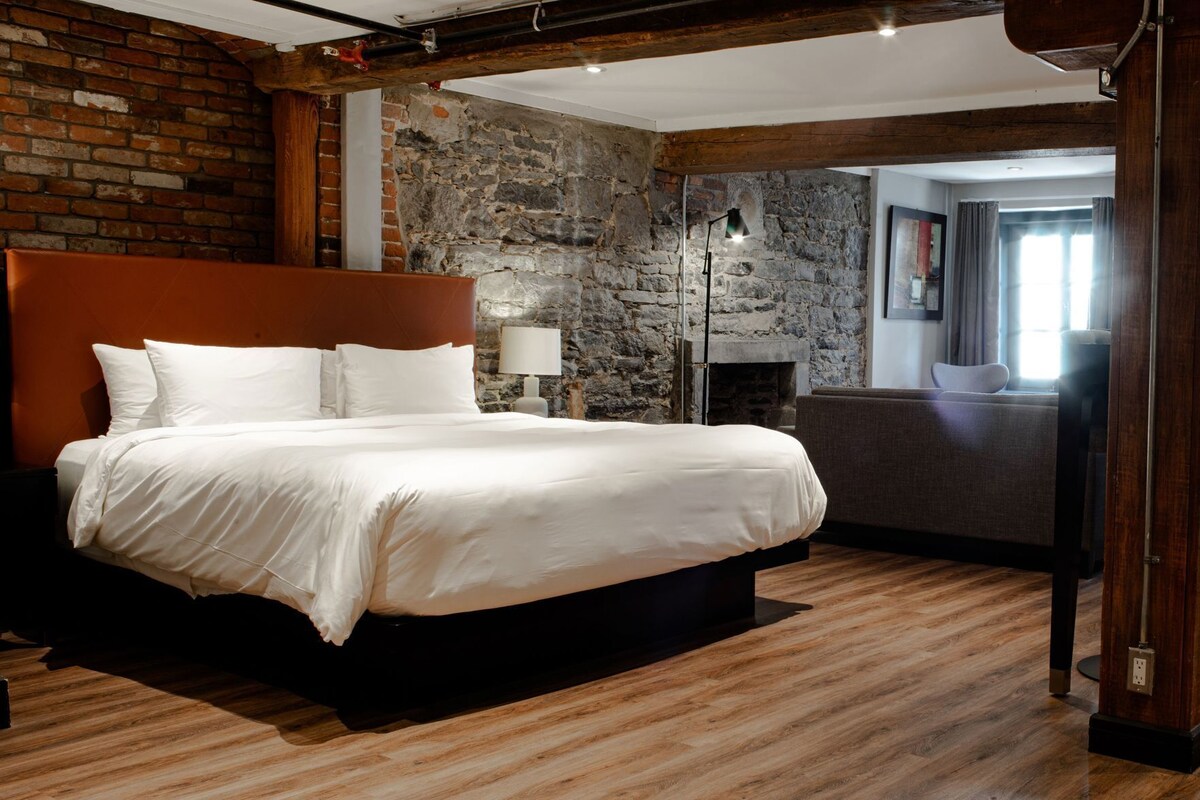 A bed in a room with a stone wall, a fireplace, and a view of a cityscape.