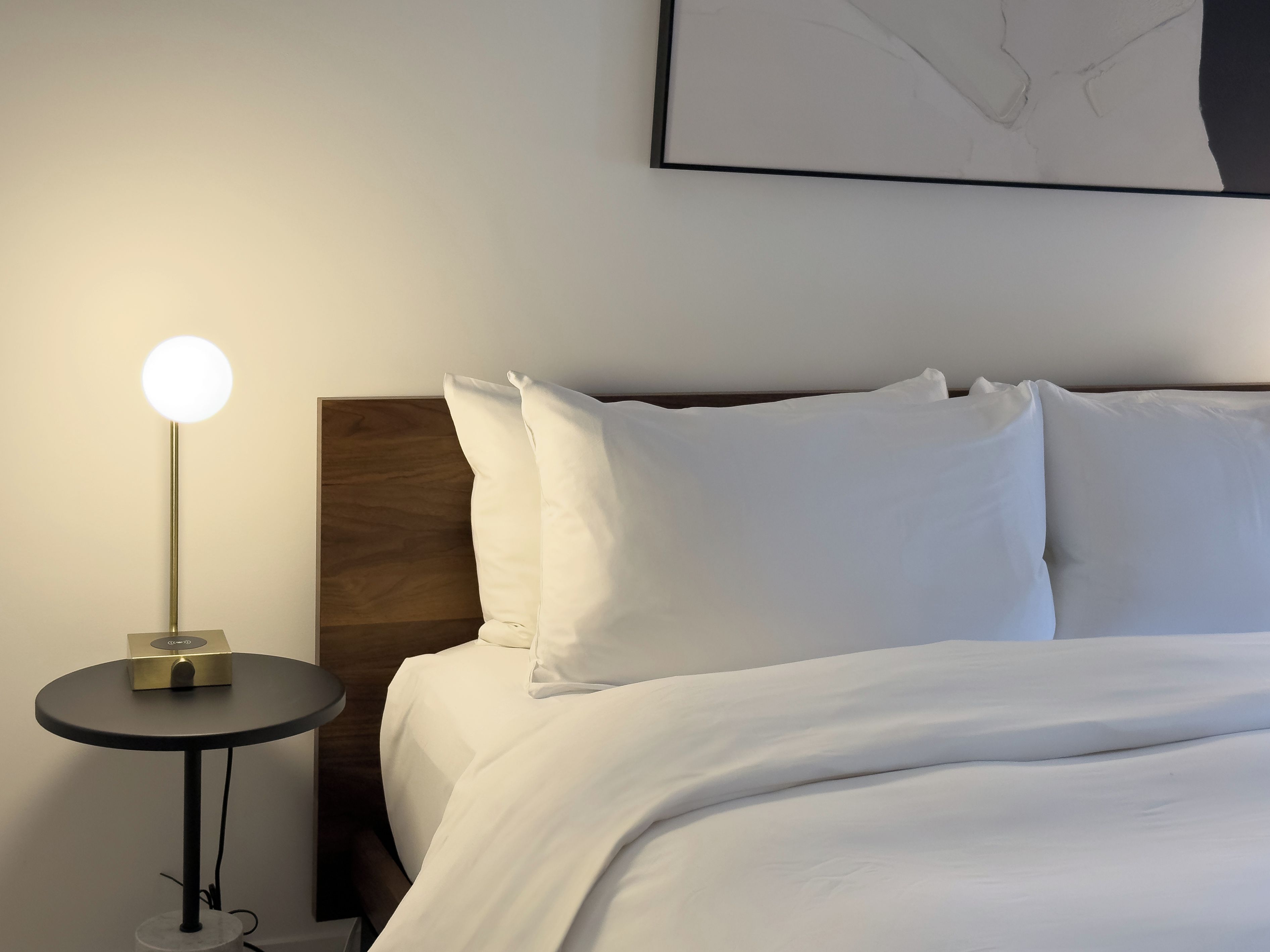 A neatly made bed with white linens, a round table with a lamp, and a framed artwork on the wall.