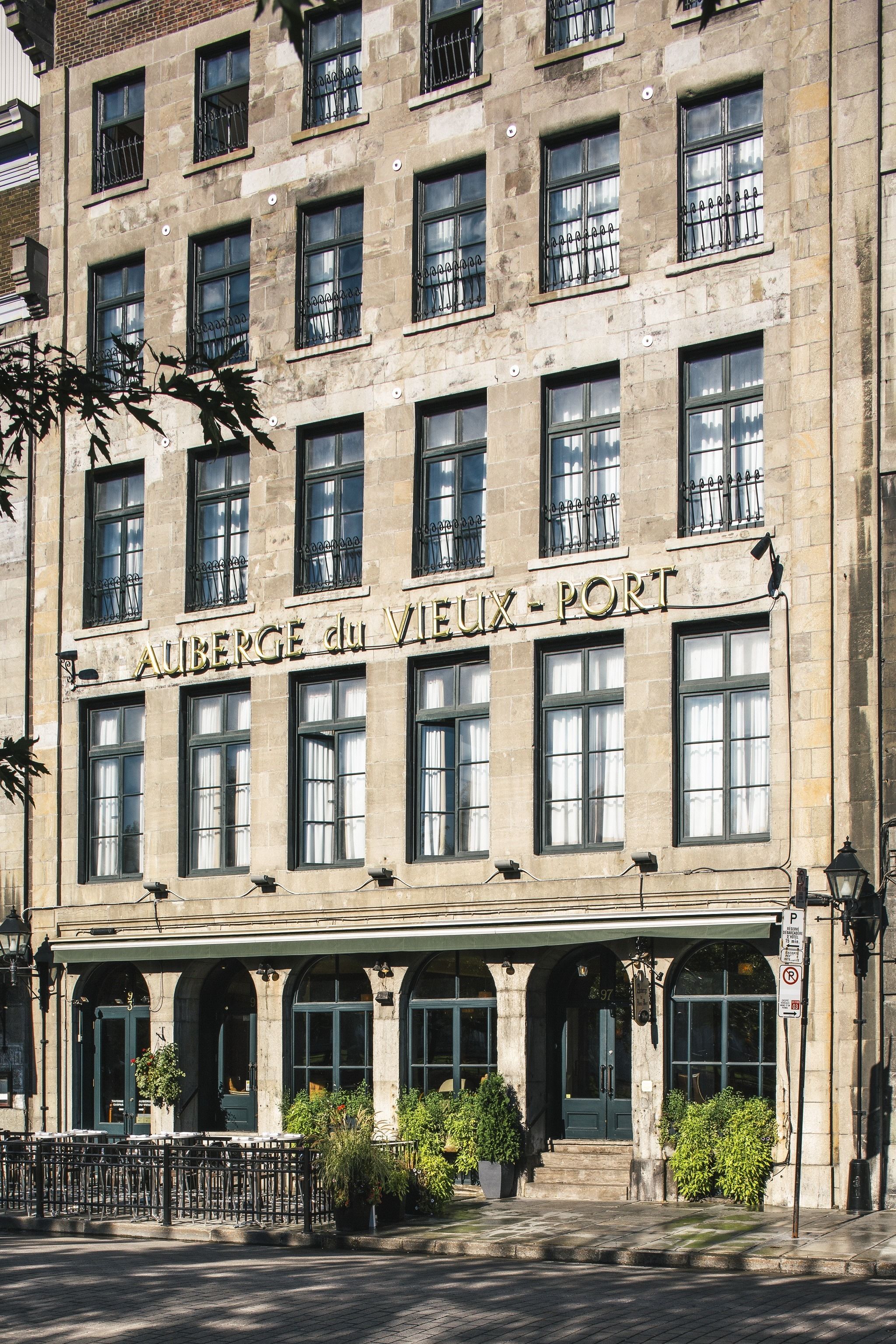 Un edificio con el letrero "AUBERGE du VIEUX - PORT". Tiene varias ventanas con marcos negros y una fachada de piedra. Hay macetas con plantas frente a la entrada.