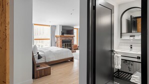 A modern hotel room with a wooden floor, a bed with a headboard, a fireplace, a large window, and a bathroom with a mirror, sink, and towel rack. - Lofts du Vieux-Port (Montreal)