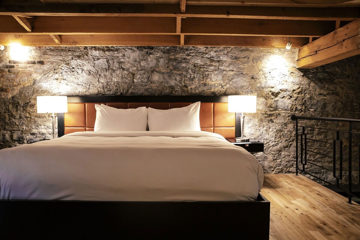 A bed in a room with a stone wall, wooden beams, and a nightstand with a lamp.