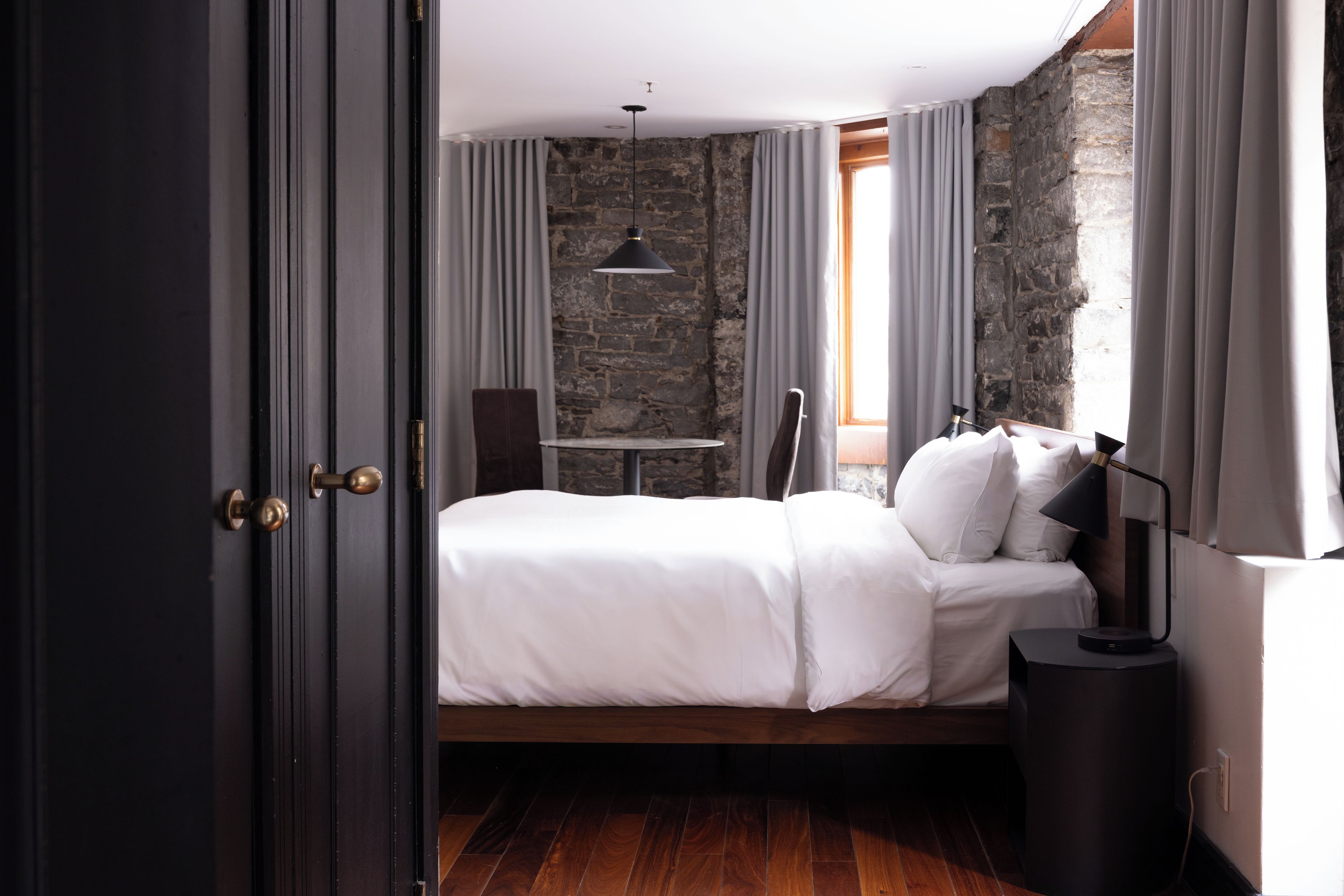A bedroom with a bed, a desk, and a chair. There is a stone wall and a window with curtains.