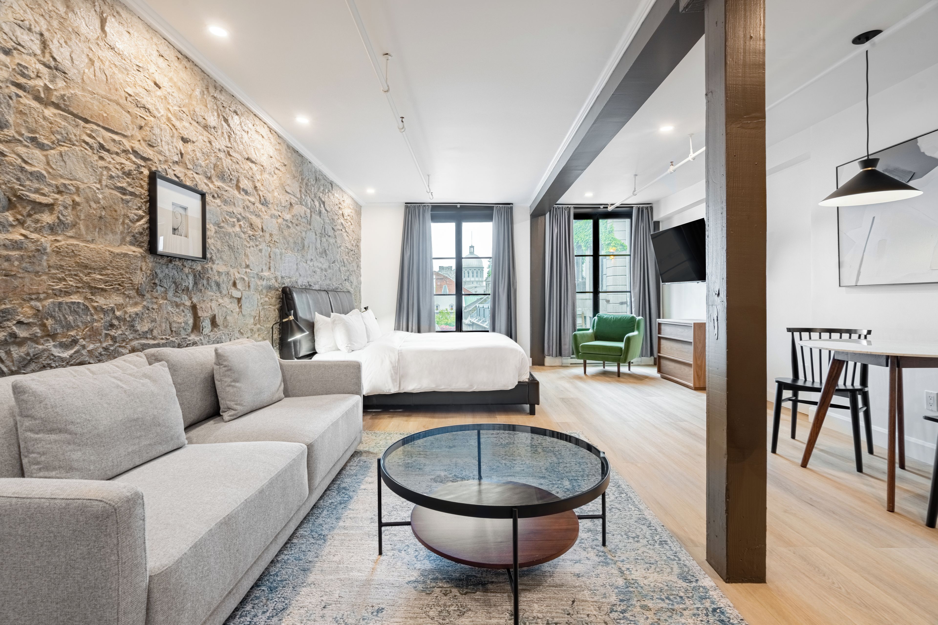 A modern bedroom with a stone wall, a bed, a round coffee table, and a view of the city.
