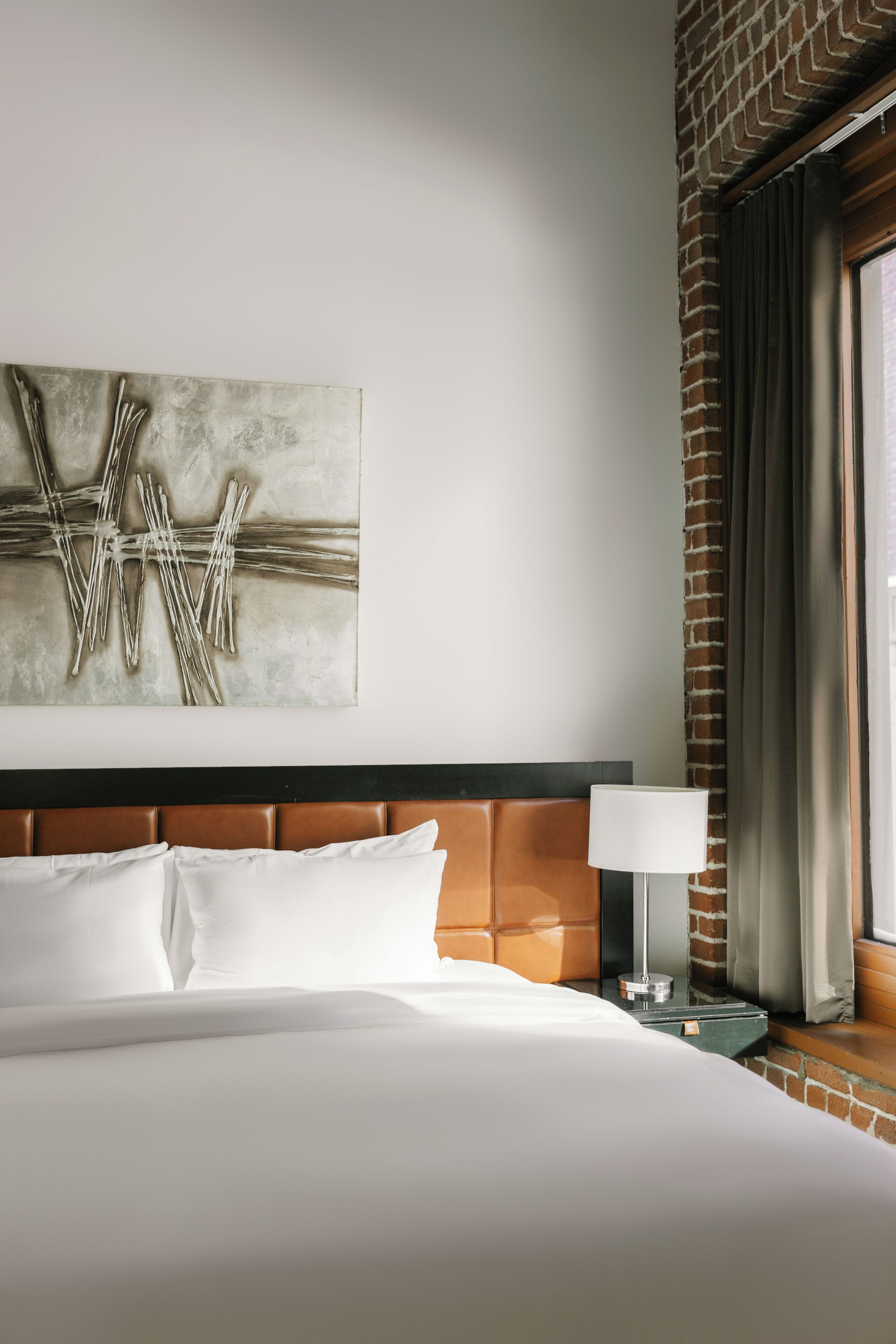 Quarto de hotel com uma cama grande, cabeceira com painel marrom, obra de arte abstrata na parede, luminária de cabeceira e uma janela com cortinas.