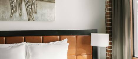 Quarto de hotel com uma cama grande, cabeceira com painel marrom, obra de arte abstrata na parede, luminária de cabeceira e uma janela com cortinas.