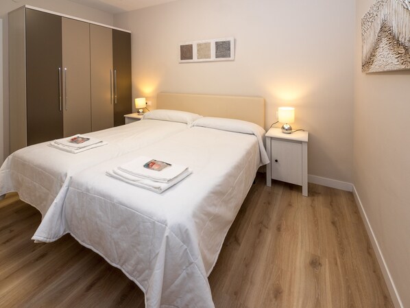 Uma cama bem arrumada com lençóis brancos e duas almofadas, com mesas de cabeceira de cada lado com abajures.