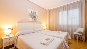 A hotel room with two beds, a nightstand with a lamp, a chair, and a window with curtains. - Gestión de Alojamientos Rooms - Guest House (Pamplona)