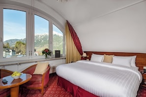 Habitación de hotel con una cama grande, zona de estar con una mesa y vistas a montañas a través de amplios ventanales.