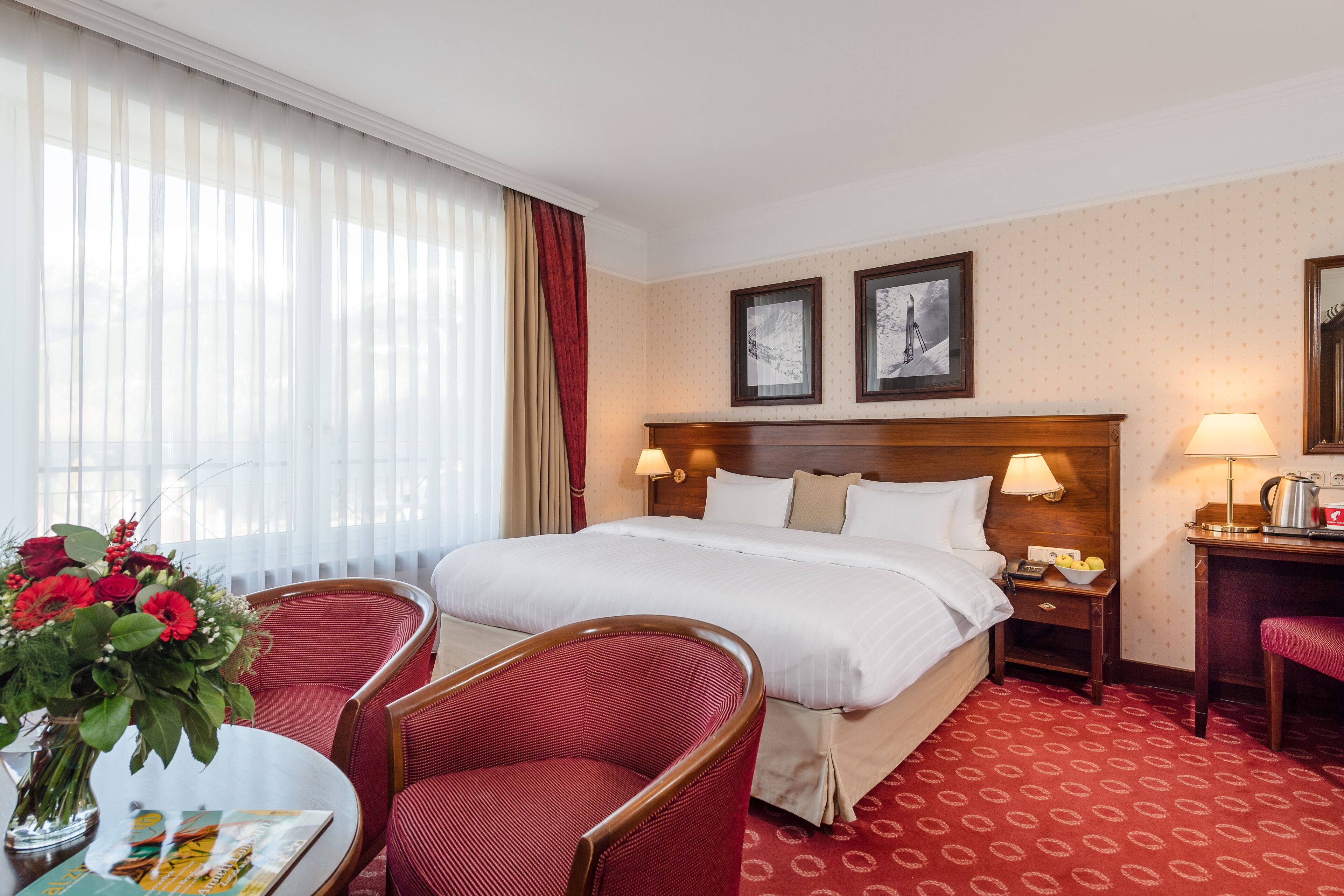 A hotel room with a large bed, two bedside lamps, a wooden nightstand, a desk with a lamp, a chair, and a vase with flowers.