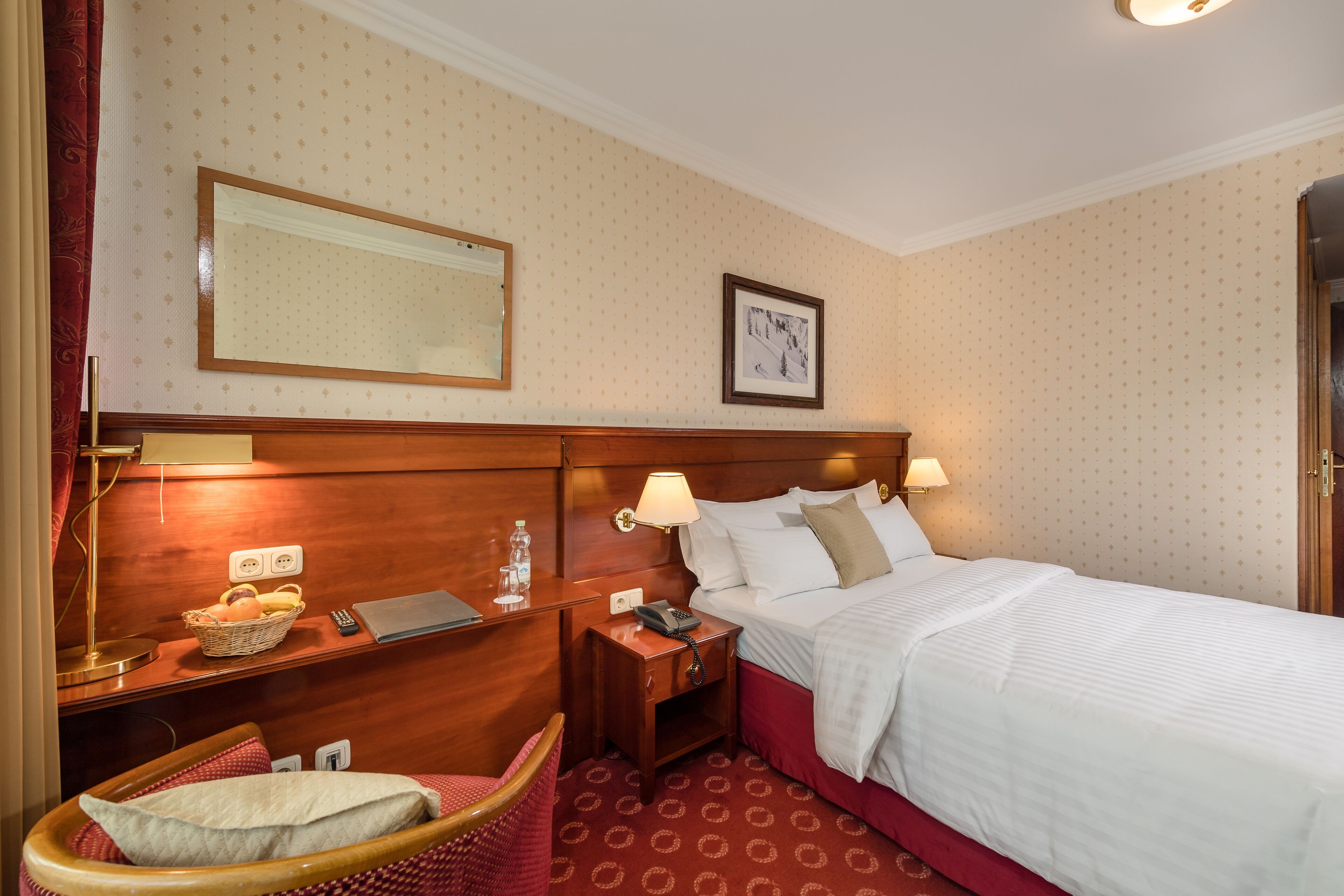 A hotel room with a bed, a nightstand, a lamp, a chair, a wall-mounted mirror, and a framed picture.