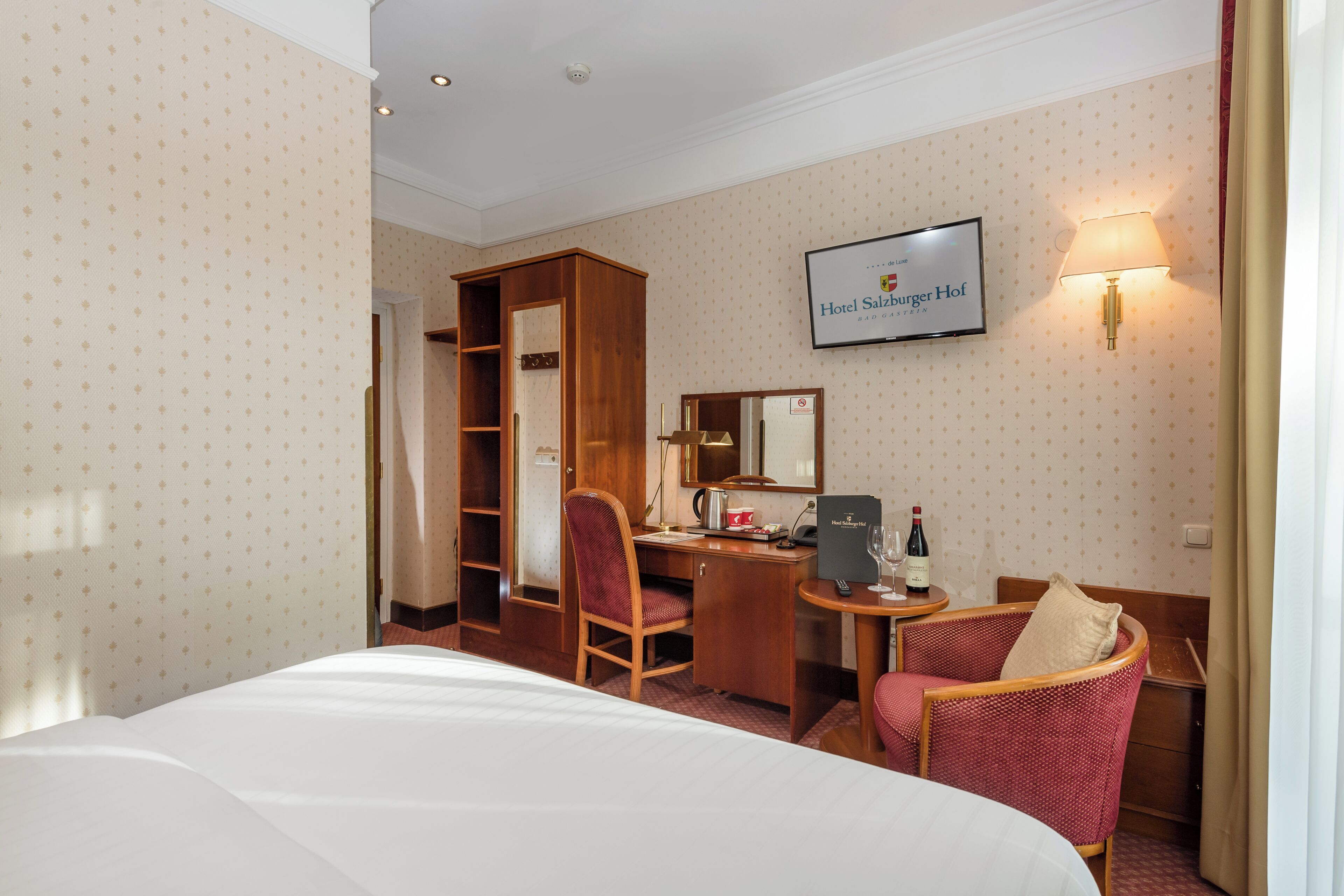 Habitación de hotel con cama, escritorio, silla, armario, espejo, televisor y lámpara.