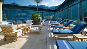 A sunlit outdoor lounge area with wooden lounge chairs and a glass enclosure, offering a view of mountains. - Hotel Salzburger Hof (Bad Gastein)