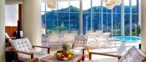 Un espacio lounge moderno con sillas blancas acolchadas, una mesa de madera con frutero, y grandes ventanales con vistas a una montaña y una piscina.
