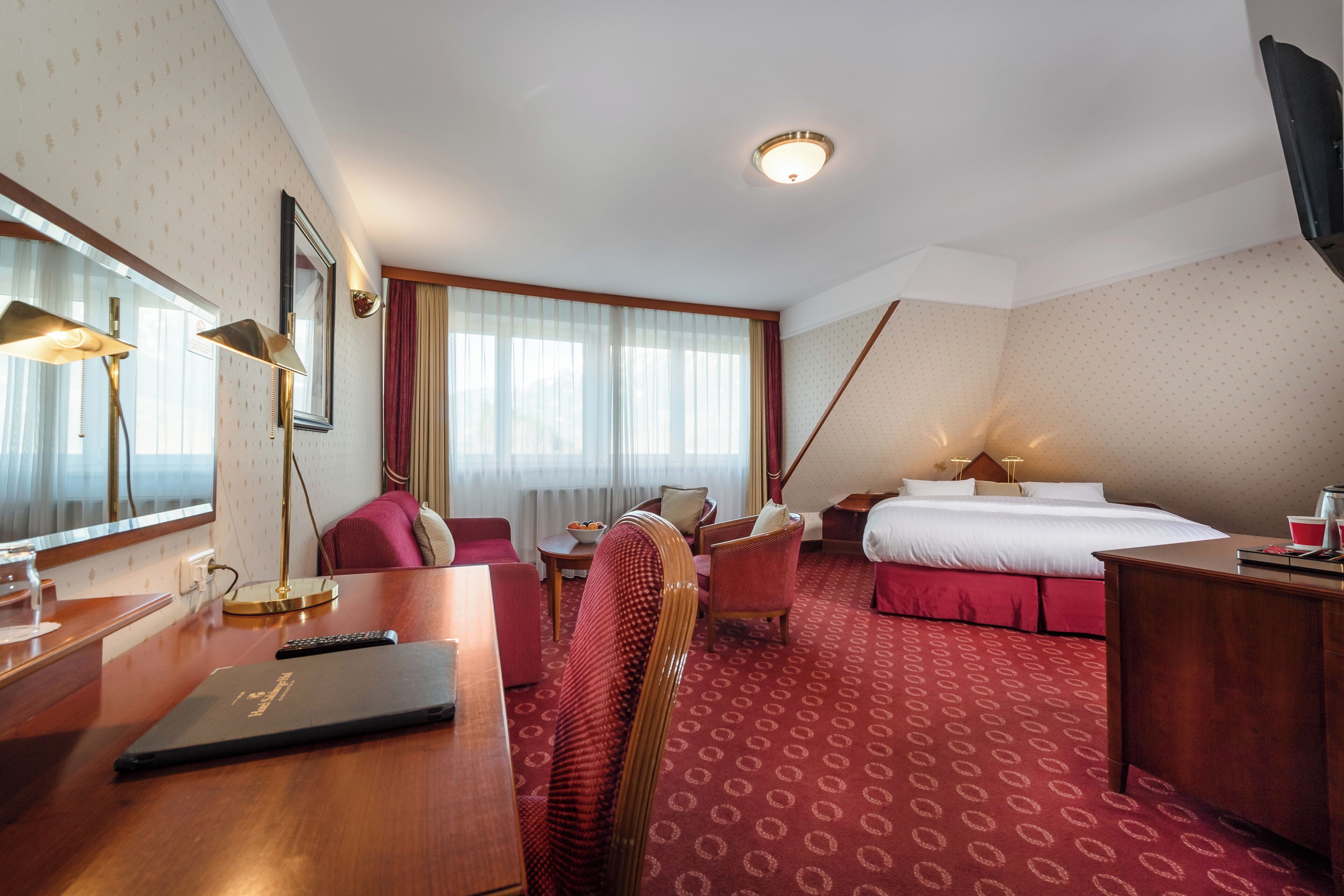 Habitación de hotel con cama, escritorio, silla, lámpara de escritorio, televisor y ventana con cortinas.