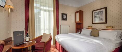 Habitación de hotel con cama, escritorio con una computadora portátil, una silla, mesita de noche, lámpara y ventana con cortinas.