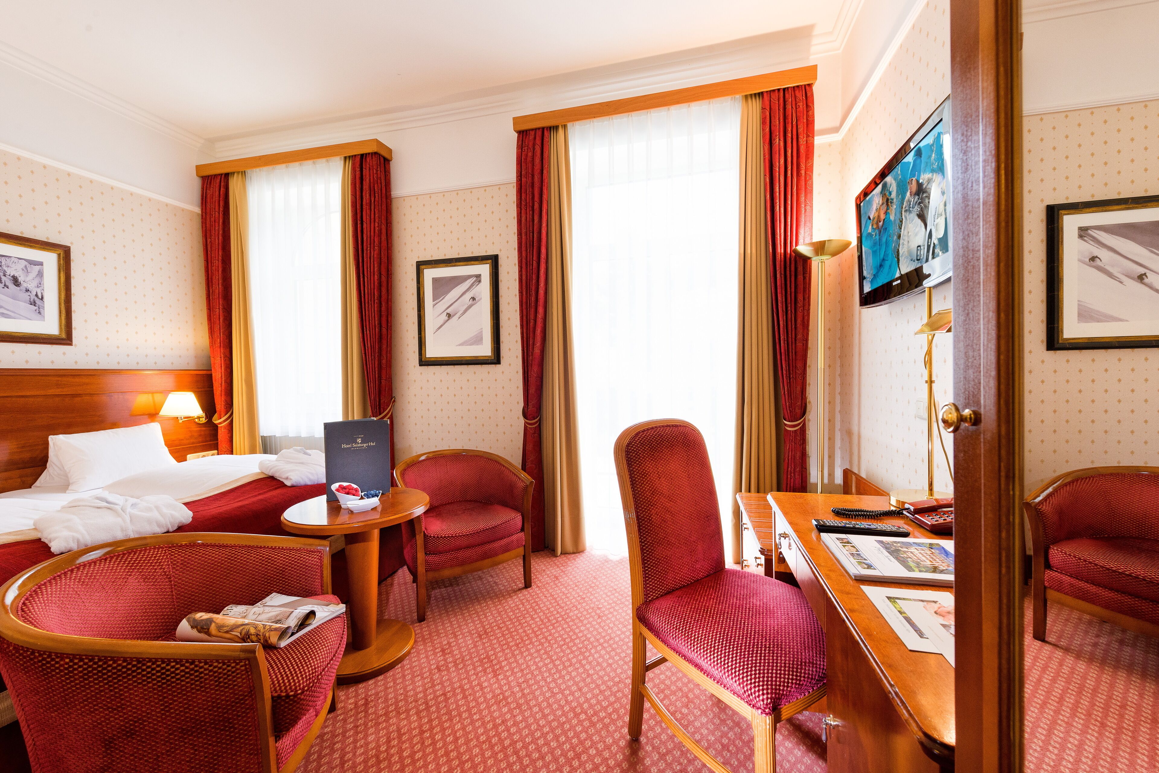 A hotel room with a bed, two red upholstered chairs, a wooden desk, a television, and framed pictures on the walls.