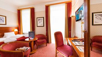 A hotel room with a bed, two red upholstered chairs, a wooden desk, a television, and framed pictures on the walls.