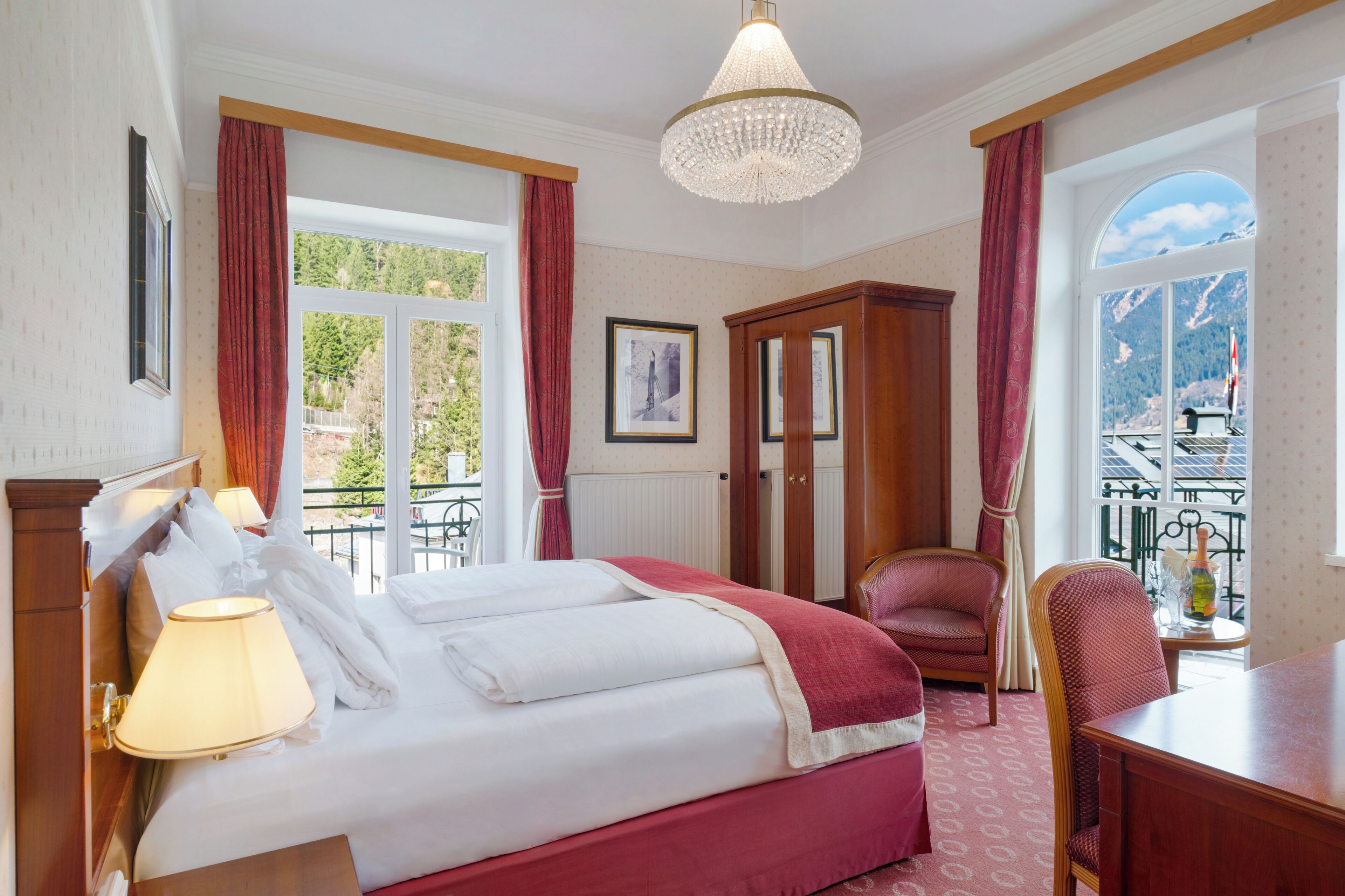 Ein Hotelzimmer mit Bett, Schreibtisch, Sessel, einer Kronleuchter und einem Fenster mit Bergblick.