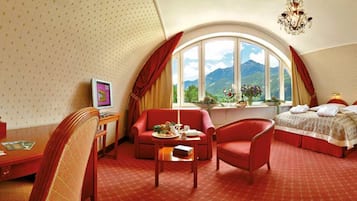 Habitación de hotel con ventanal curvo, cama, escritorio, silla y una mesita con un jarrón de flores.