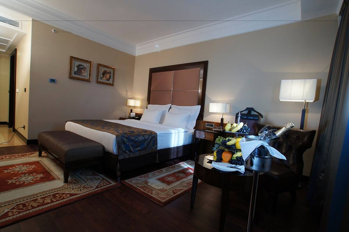 A hotel room with a bed, two bedside tables, a chair, a desk with fruit and a lamp, and a red and beige carpet.