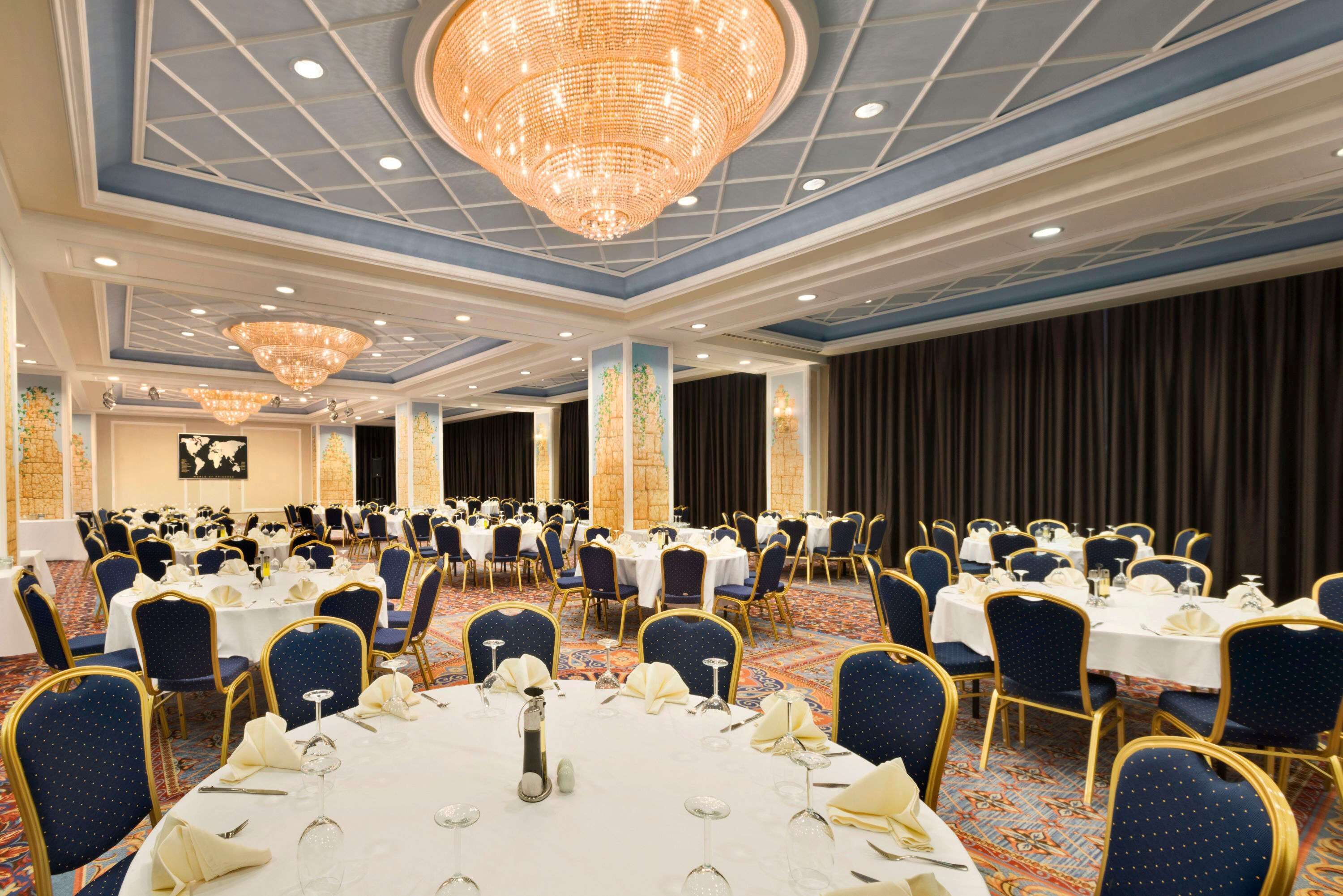 A banquet hall with round tables set for dining, blue upholstered chairs, and a large chandelier.