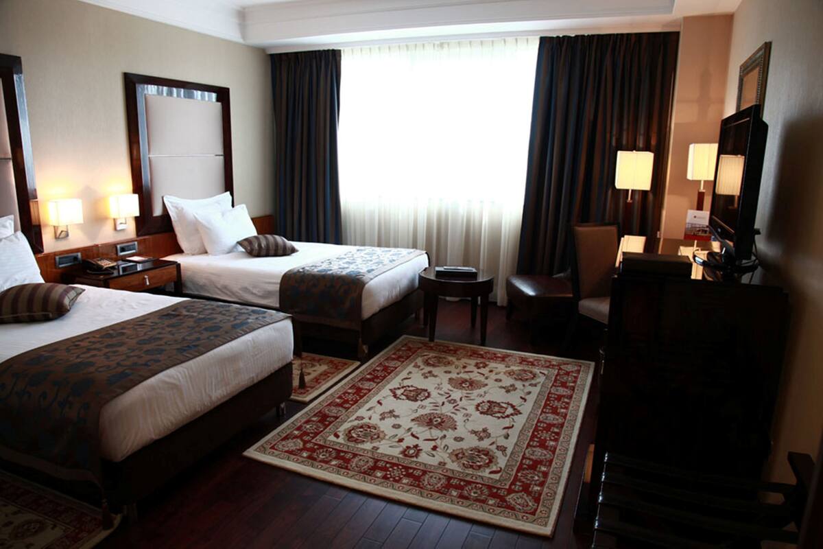 A hotel room with two beds, a desk, a chair, a TV, and a large window with curtains.