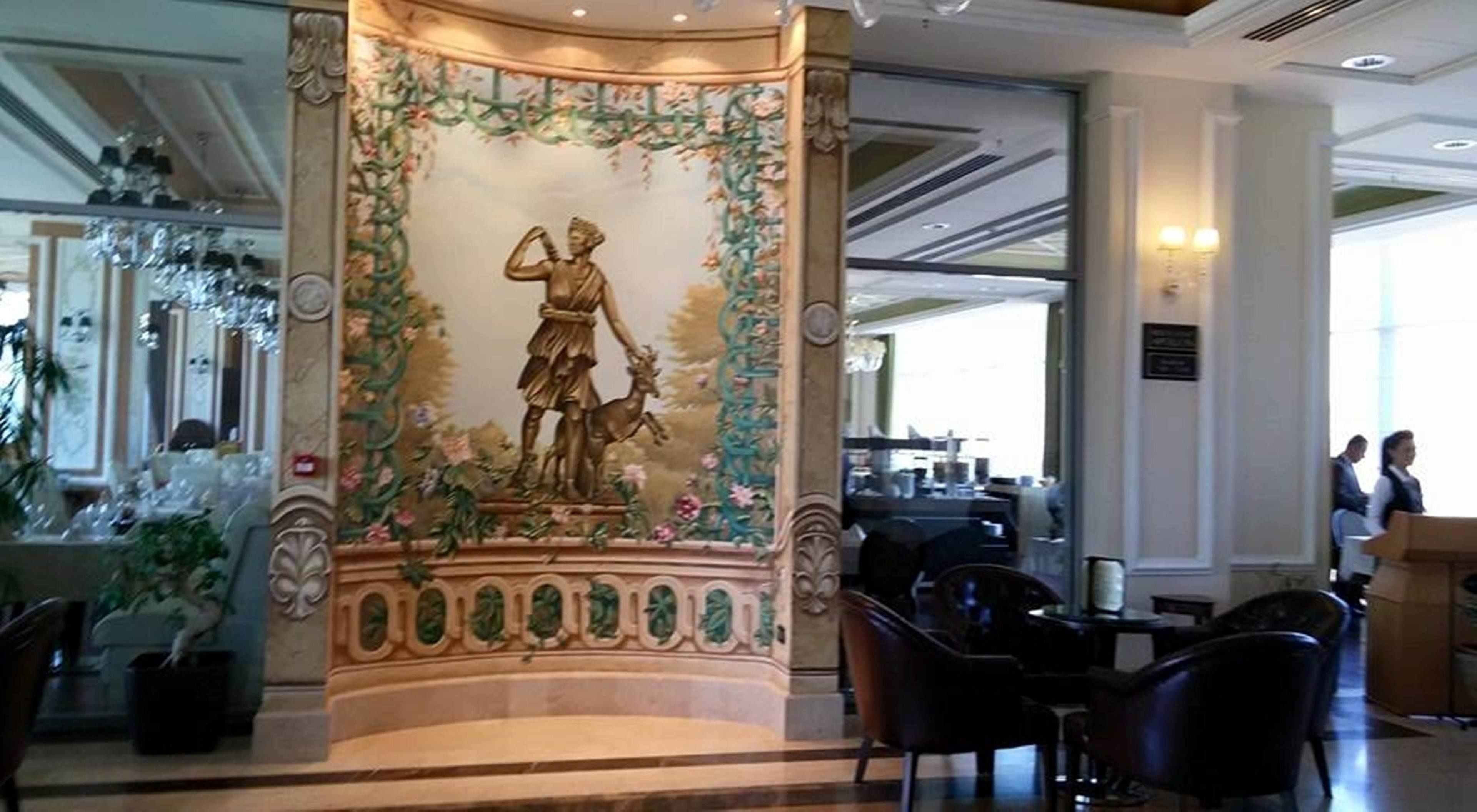 Un hall d’entrée doté d’une colonne centrale décorative surmontée d’une statue, entouré d’espaces de détente et d’un bar à café.