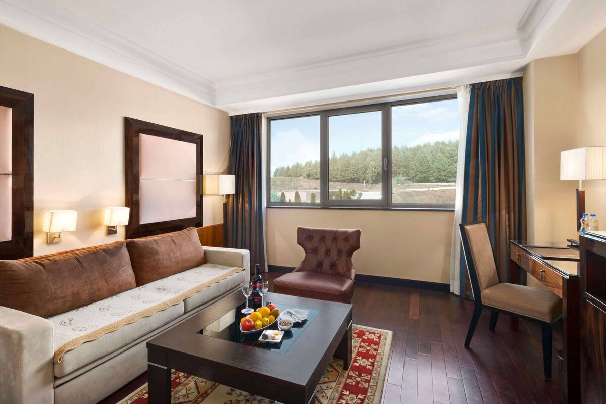 A hotel room with a sofa, armchair, dining table, and a large window offering a view of trees.