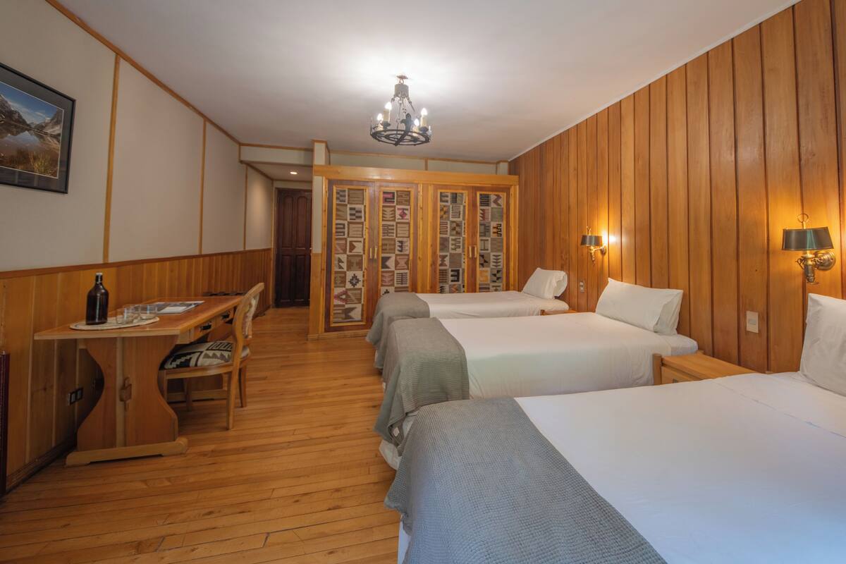 A hotel room with wooden paneling, two beds, a desk with a bottle and glasses, a chandelier, and a wall with patterned tiles.