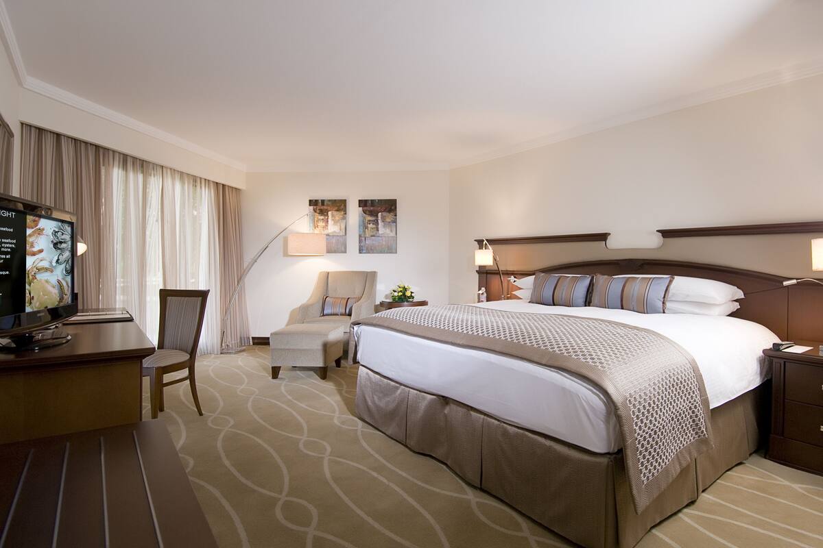 A hotel room with a large bed, a desk with a television, a chair, and a lamp.