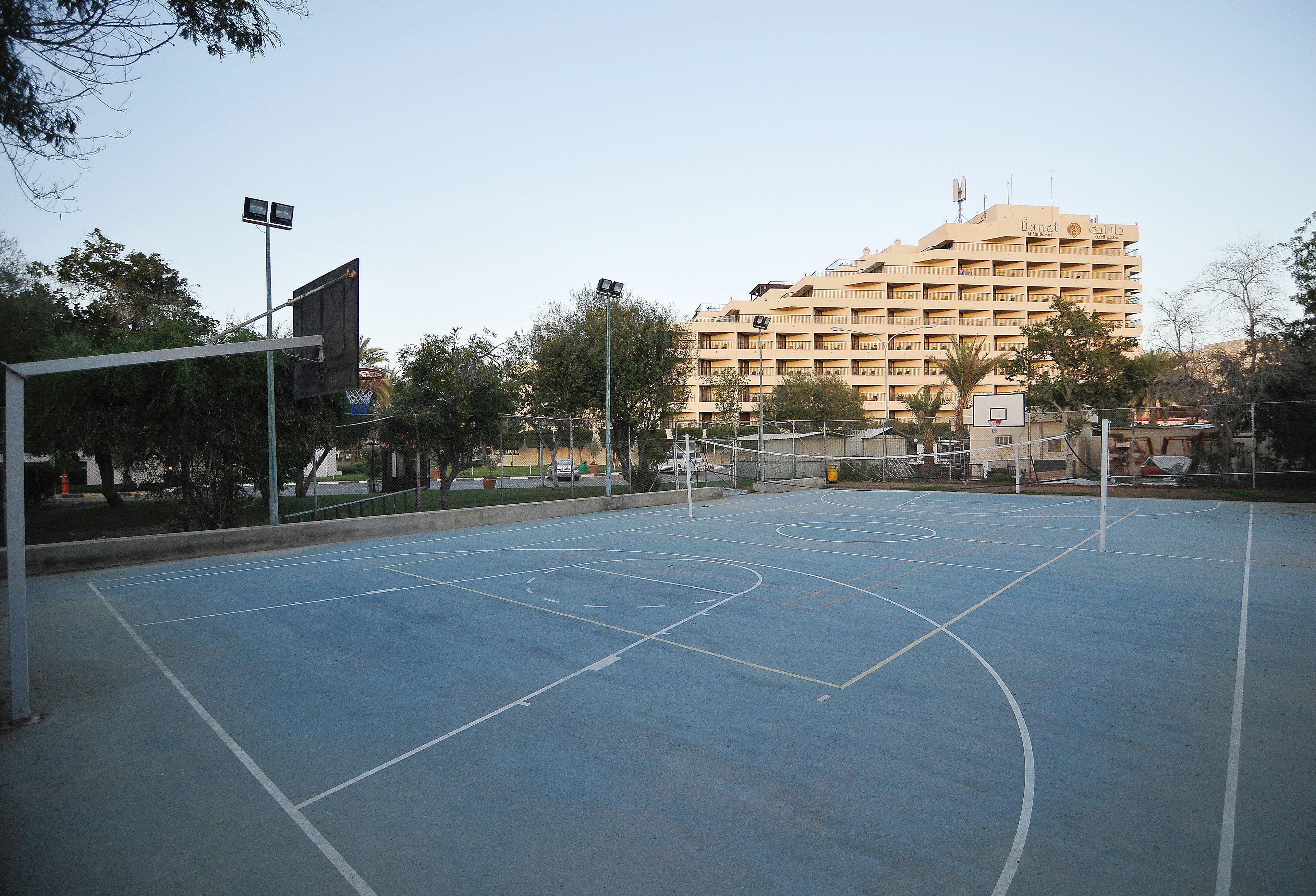 An outdoor basketball court with a net, surrounded by trees and a multi-story building in the background.