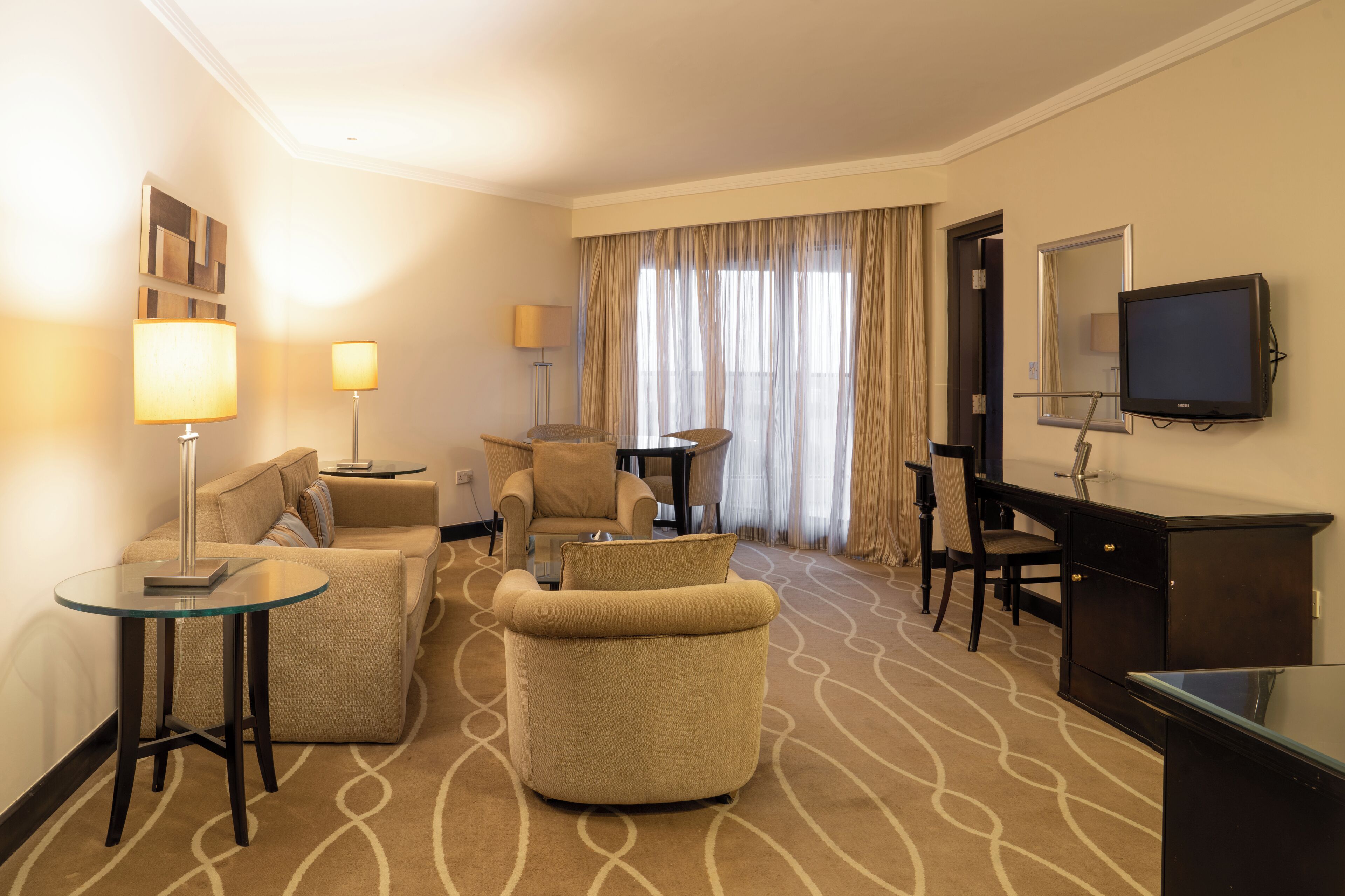 Habitació d'hotel amb un sofà, butaques, un escriptori, un televisor i un sòl amb moqueta.