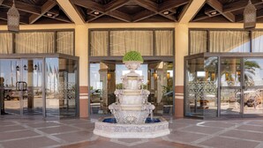Una fuente de varios niveles con un topiario verde en el centro, rodeada por una base decorativa, ubicada en un patio con piso de baldosas y puertas de cristal que dan a un edificio con techo de madera y ventanas amplias.