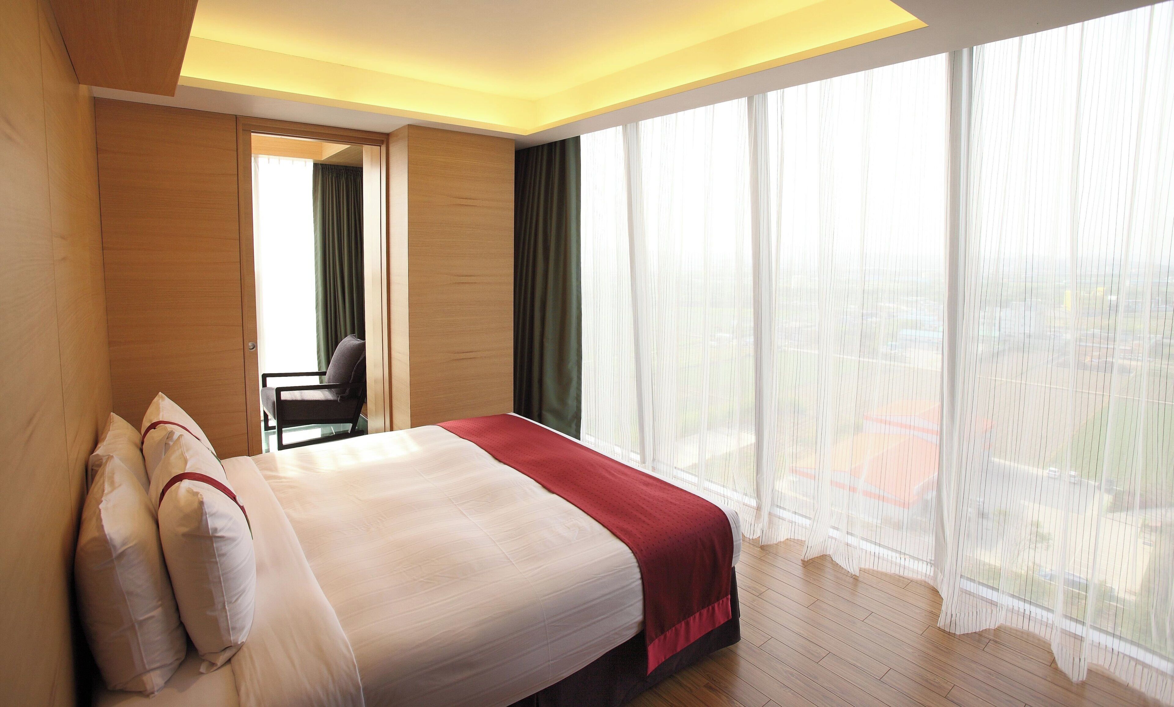 A hotel room with a bed, a chair, a desk, and large windows with sheer curtains.