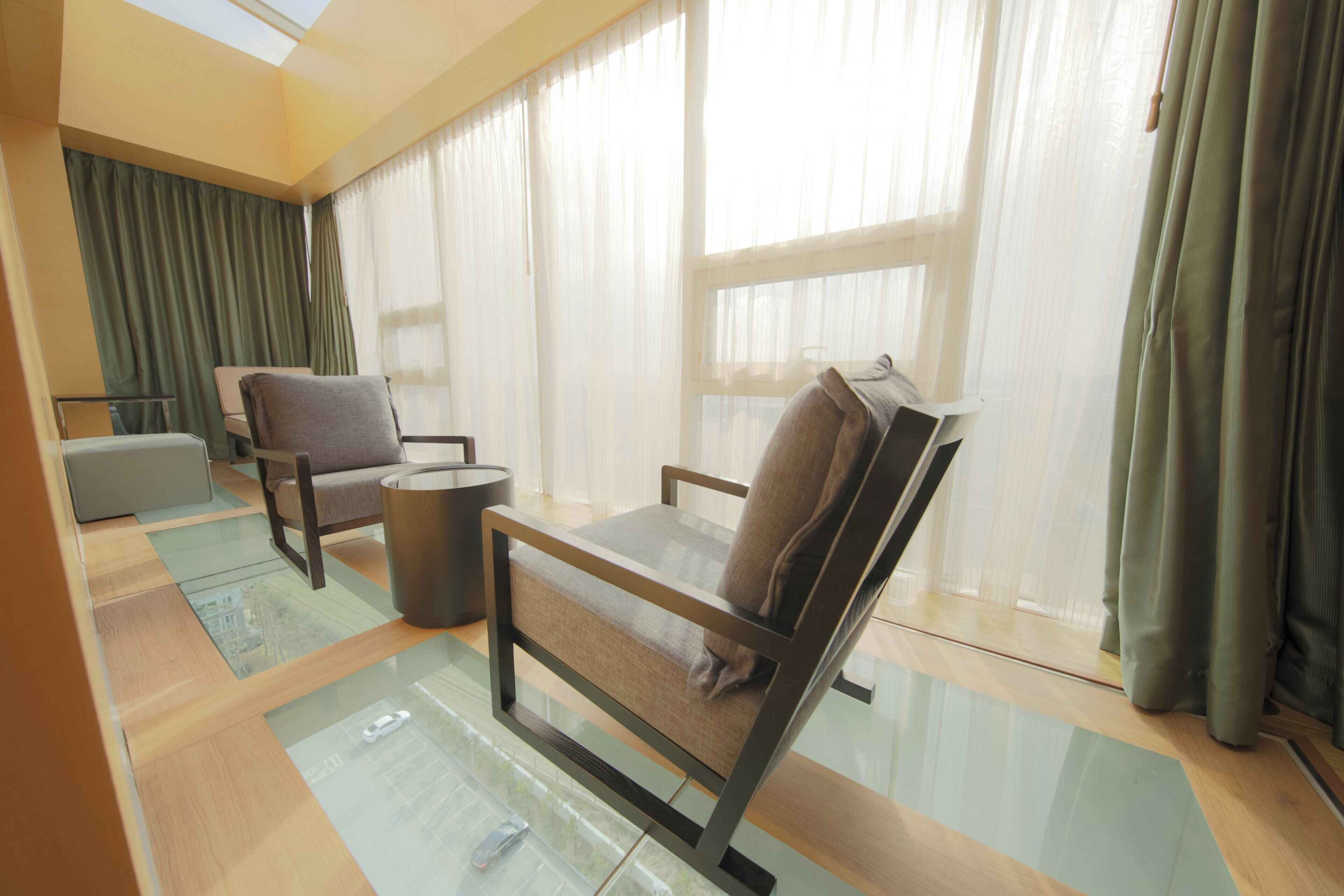 A modern living room with a glass coffee table, two armchairs, a sofa, and sheer curtains.