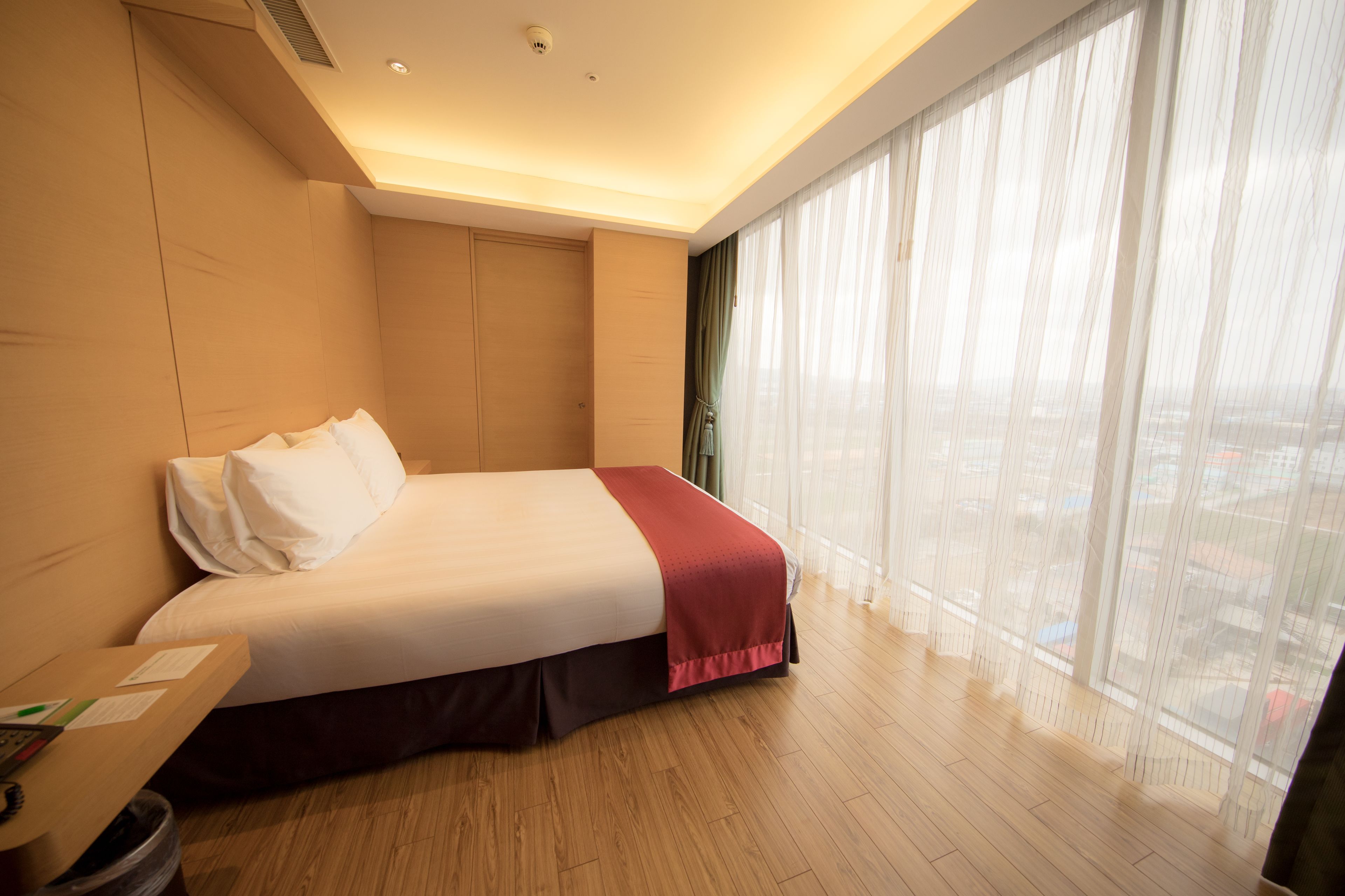 A hotel room with a bed, a desk, and a large window with sheer curtains.