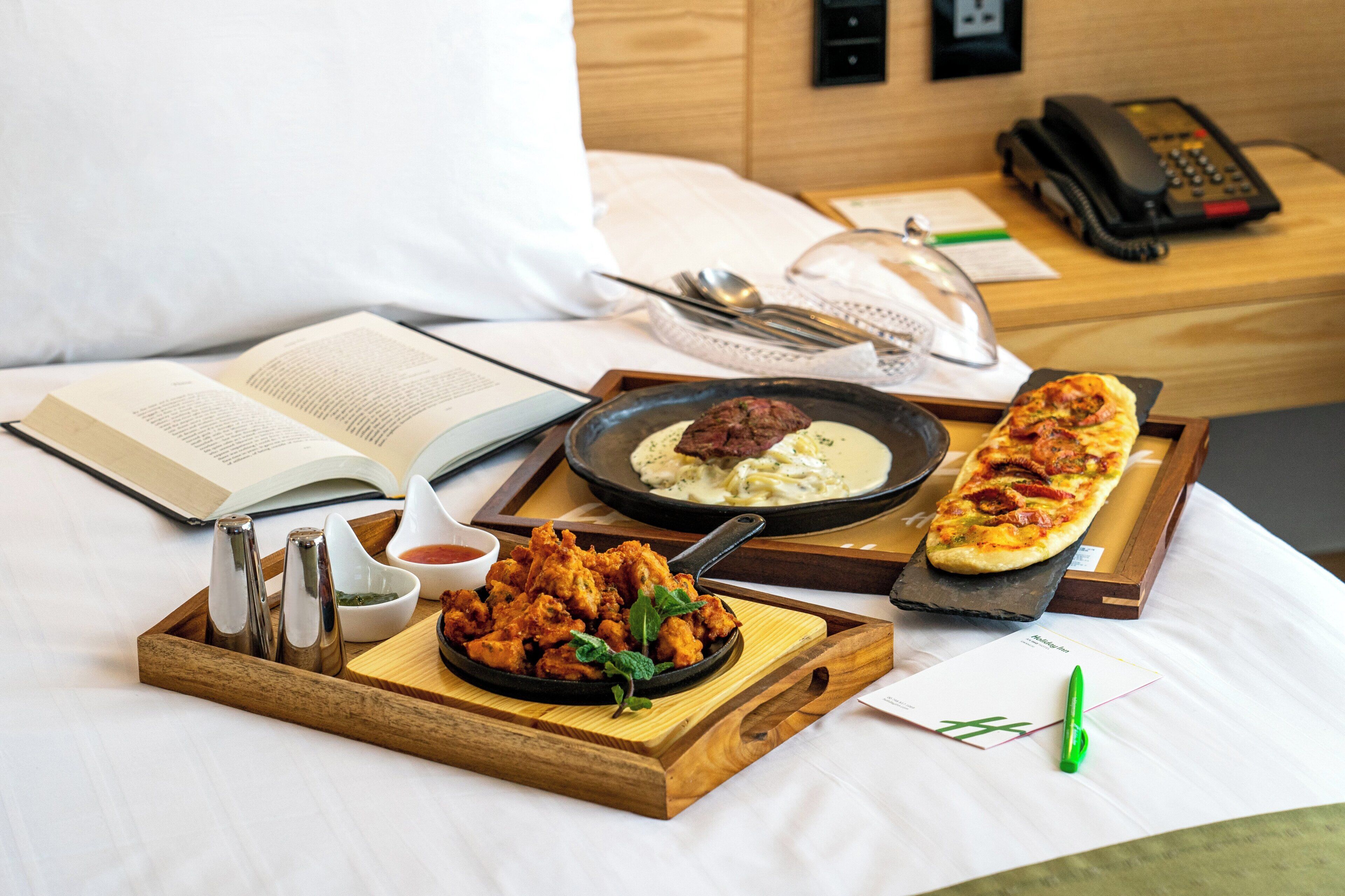 A neatly arranged breakfast tray with a variety of dishes, a book, and a remote control on a bed.