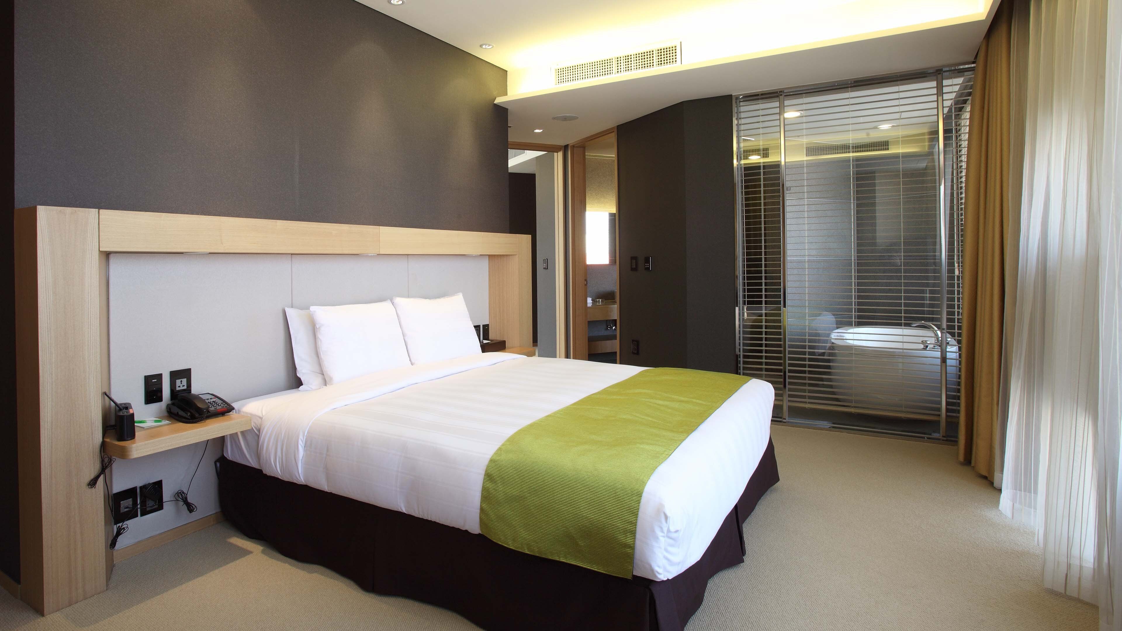 A hotel room with a large bed, a bedside table with a phone and charger, a walk-in closet, and a sliding glass door leading to a bathroom.